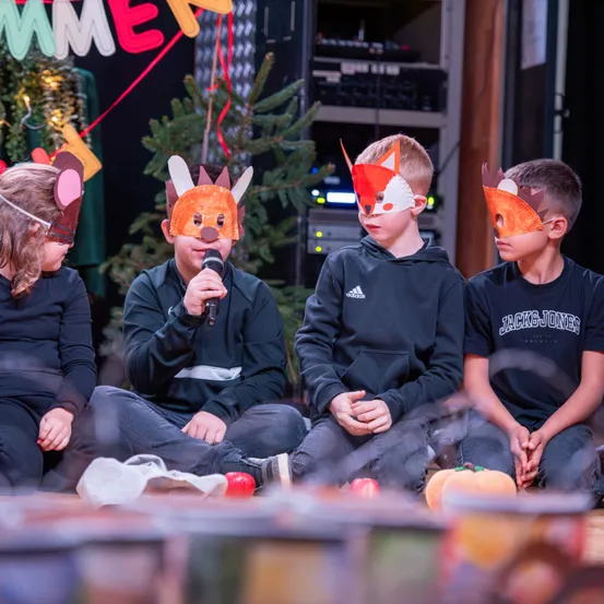 Vier Kinder sitzen auf dem Boden, jedes trägt eine Fuchsmaske. Eines hält ein Mikrofon, ein anderes einen Apfel. Sie sind in einem Raum mit einem Weihnachtsbaum und einem Banner.