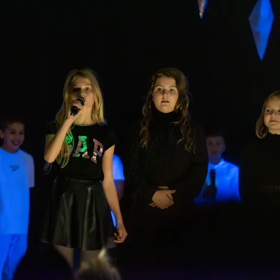 Drei junge Mädchen treten auf der Bühne unter blauem Licht auf. Das Mädchen in der Mitte hält ein Mikrofon und singt. Hinter ihr hat ein anderes Mädchen die Hände in die Taille gestemmt. Ein Junge im Hintergrund beobachtet sie.