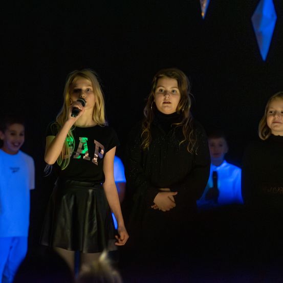 Drei junge Mädchen treten auf der Bühne unter blauem Licht auf. Das Mädchen in der Mitte hält ein Mikrofon und singt. Hinter ihr hat ein anderes Mädchen die Hände in die Taille gestemmt. Ein Junge im Hintergrund beobachtet sie.