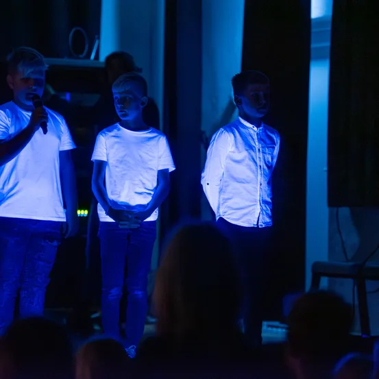 Drei Jungen stehen auf einer Bühne, beleuchtet von blauen Lichtern, und führen ein Programm vor einem Publikum auf. Ein Junge hält ein Mikrofon.