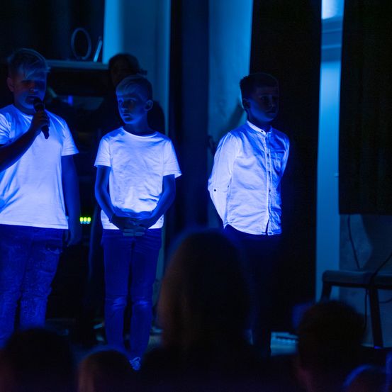 Drei Jungen stehen auf einer Bühne, beleuchtet von blauen Lichtern, und führen ein Programm vor einem Publikum auf. Ein Junge hält ein Mikrofon.