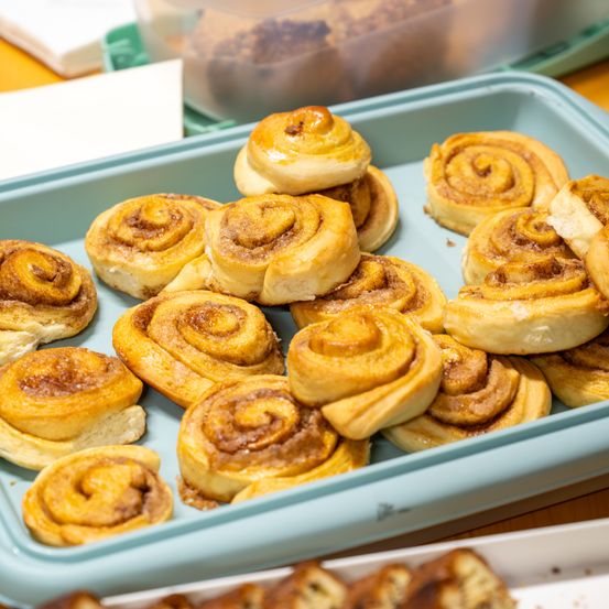 Ein Tablett gefüllt mit goldenbraunen Zimtschnecken. Das Tablett ist blau, und es gibt einige Krümel auf dem Tablett. Im Hintergrund befinden sich weitere Tabletts mit Backwaren.