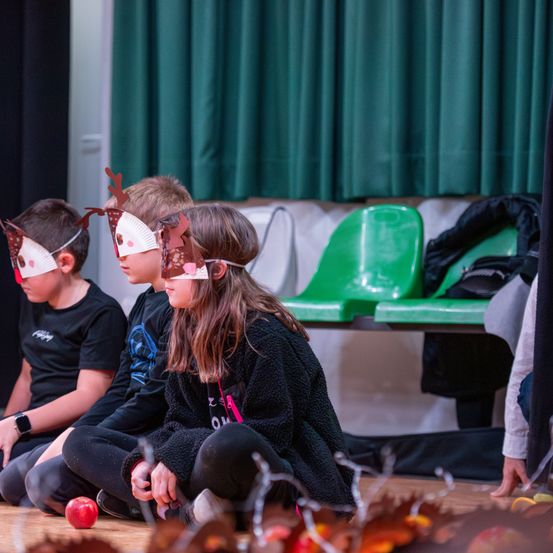 Drei Kinder mit Papier-Rehmasken sitzen auf dem Boden und beobachten wahrscheinlich eine Aufführung, mit einem grünen Stuhl und einem Vorhang im Hintergrund.