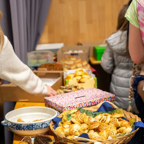 Ein Tisch mit verschiedenen Gebäckstücken, darunter Croissants und Brot, steht in einem Raum mit Holzboden. Menschen sind in der Nähe, und einer greift nach den Croissants.