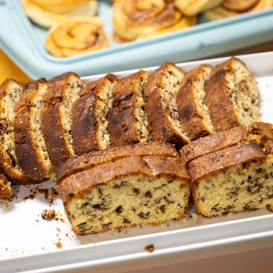 Ein weißer Teller mit geschnittenem Schoko-Bananen-Kuchen, im Hintergrund befinden sich Zimtschnecken in einer blauen Schale.