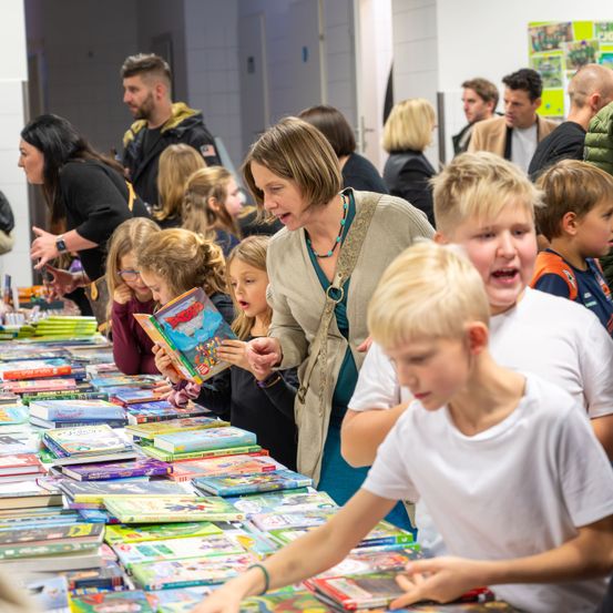 Eine Gruppe von Kindern und Erwachsenen versammelt sich um einen mit Büchern gefüllten Tisch und betrachtet sie. Eine Frau in der Mitte hält ein Buch und spricht mit den Kindern.