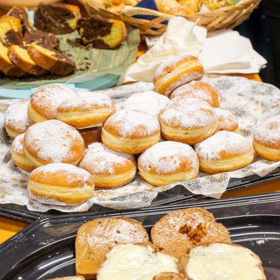 Ein Tisch mit verschiedenen Gebäcksorten, darunter mit Puderzucker bestreute Donuts und andere Backwaren wie Brotscheiben, alles auf einem blumengemusterten Tuch angeordnet.