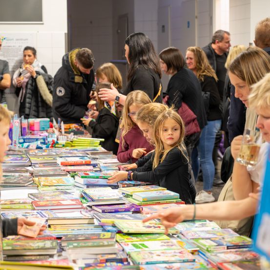 Eine Gruppe von Menschen, darunter Familien mit Kindern, versammelt sich um einen Tisch voller bunter Bücher. Sie scheinen auf einem Büchermarkt oder einer Buchverkaufsveranstaltung zu sein.