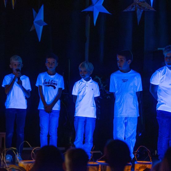 Fünf Jungen stehen auf einer Bühne vor einem Publikum. Sie tragen alle weiße Hemden und blaue Hosen. Einer der Jungen hält ein Mikrofon. Über ihnen hängen weiße Sterne von der Decke.