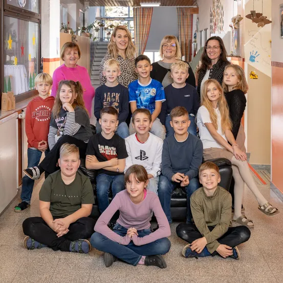 Eine Gruppe von Kindern und Erwachsenen, darunter Frauen, posiert für ein Foto in einem Flur. Der Flur hat eine Treppe und ein Fenster.