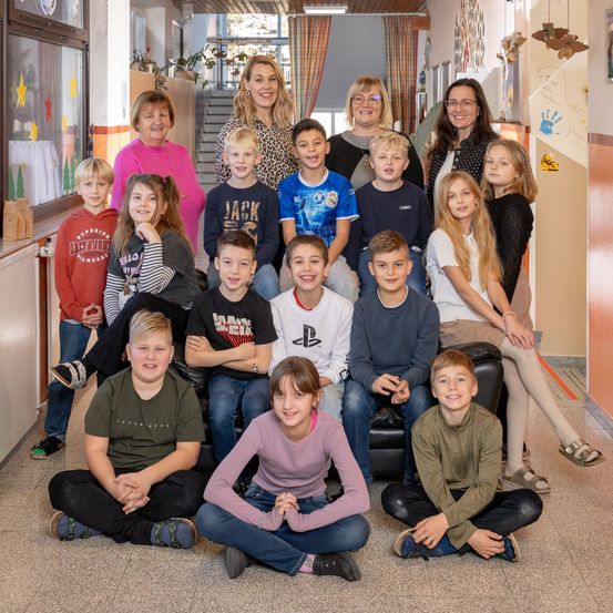 Eine Gruppe von Kindern und Erwachsenen, darunter Frauen, posiert für ein Foto in einem Flur. Der Flur hat eine Treppe und ein Fenster.