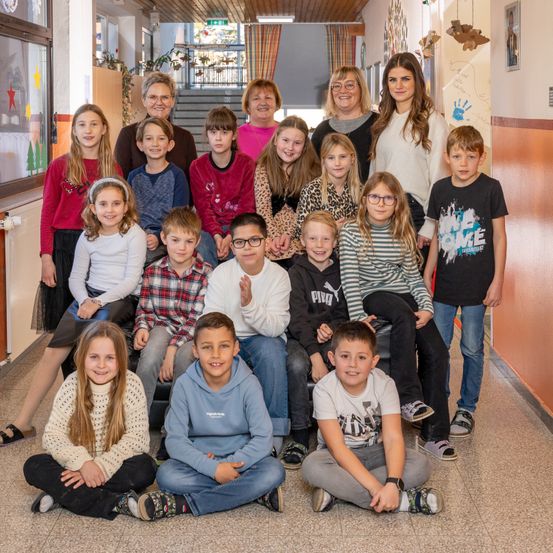 Eine Gruppe von Kindern und Erwachsenen, einschließlich einer Lehrerin, posiert für ein Foto in einem Flur. Die Kinder sitzen auf dem Boden und lächeln.