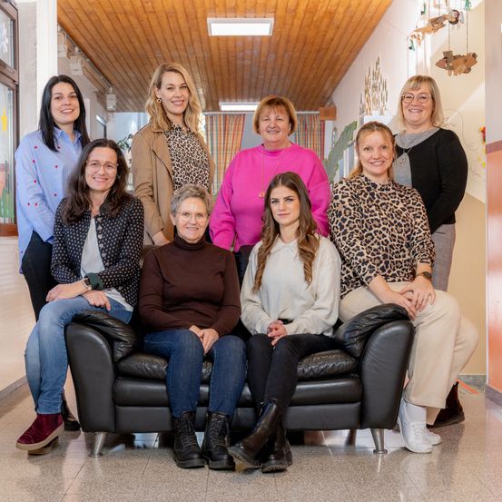 Eine Gruppe von Frauen posiert für ein Foto in einem Flur. Sie stehen alle lächelnd hinter einer schwarzen Couch. Die Frau in der Mitte sitzt auf der Couch. Der Flur hat Holzdecken und Wände mit Dekorationen.