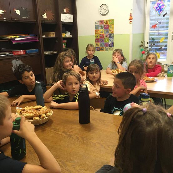 Ein Klassenzimmer mit Kindern beim Essen. Eine Uhr ist an der Wand. Ein Kind isst mit den Händen, und andere sitzen an Tischen mit Essen.