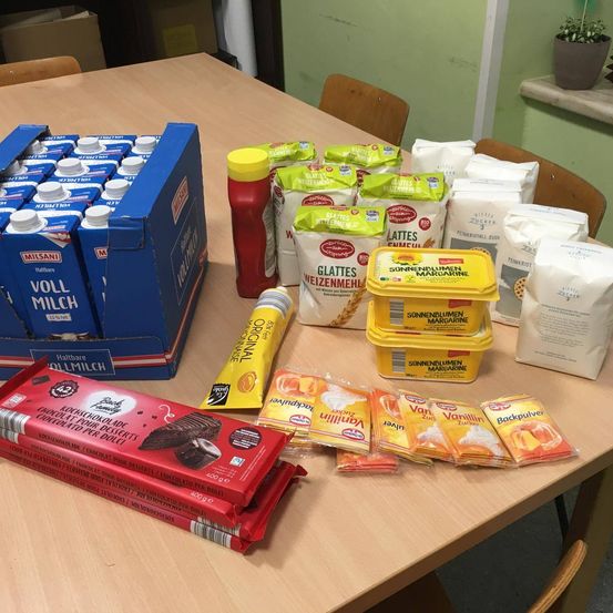 Auf einem Holztisch sind eine blaue Kartonschachtel mit Milchkartons, rote Soße, gelbe Margarine, Mehl und andere Lebensmittel zu sehen. Eine rote Schokoladentafel ist ebenfalls vorhanden.