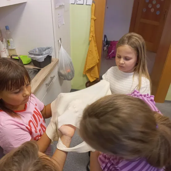 Einige Mädchen arbeiten in einem Raum an einem Projekt. Eines der Mädchen hält ein weißes Tuch und ein anderes schaut es sich an. Ein Mülleimer steht auf der Arbeitsplatte, und ein gelber Handtuch hängt an der Wand.