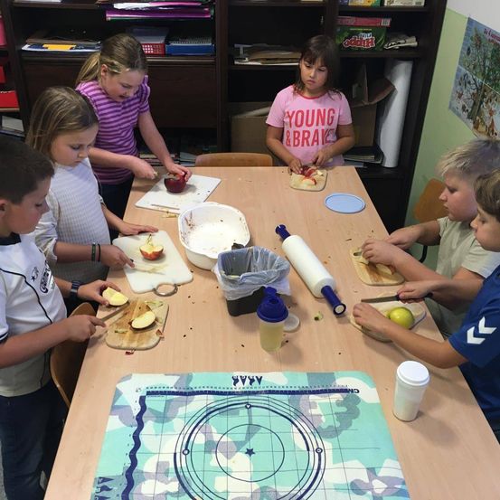 Kinder versammeln sich um einen Tisch, jedes Kind hält ein Messer und schneidet Äpfel. Eines der Mädchen trägt ein rosa Shirt mit der Aufschrift 'Young & Brave'.