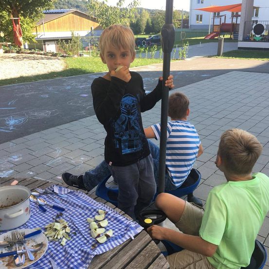Drei Kinder sitzen um einen Tisch mit Essen, eines isst einen Apfel, in einem Außenbereich.