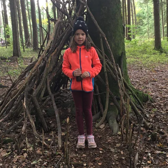 Ein junges Mädchen steht vor einer großen Struktur aus Ästen im Wald. Sie lächelt.