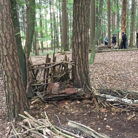 Eine Gruppe von Menschen befindet sich in einem Wald mit einer behelfsmäßigen Unterkunft aus Stöcken und Steinen. Einige tragen Mützen.