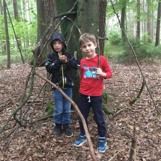 Zwei Jungen stehen im Wald, einer hält einen Stock. Sie sind von Stöcken und Blättern umgeben und lächeln für ein Foto.