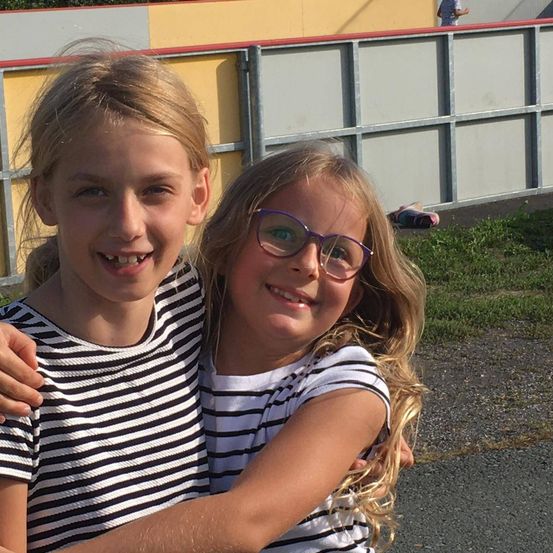 Zwei Mädchen, eines mit Brille und das andere lächelnd, stehen eng beieinander und umarmen sich. Der Hintergrund zeigt einen Zaun und einen Rasenbereich.