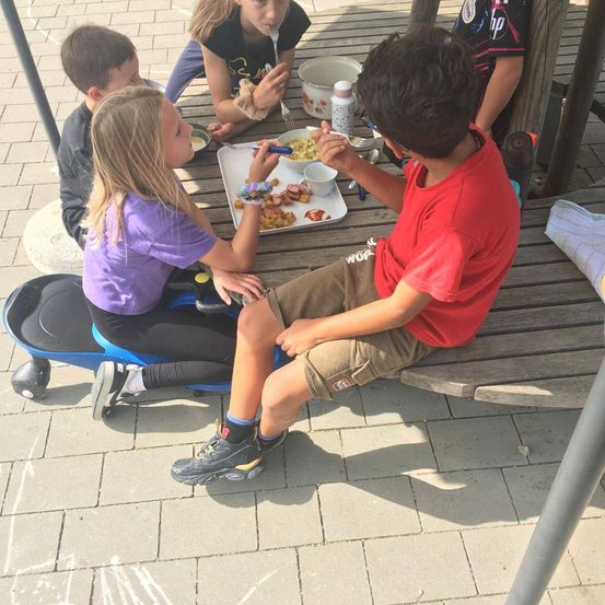 Eine Gruppe von Kindern sitzt auf einer Bank und isst. Ein Mädchen sitzt auf einem Roller und ein Junge neben ihr. Sie essen mit Löffel und Gabel. Ein anderes Mädchen sitzt auf der Bank und hält eine Flasche.