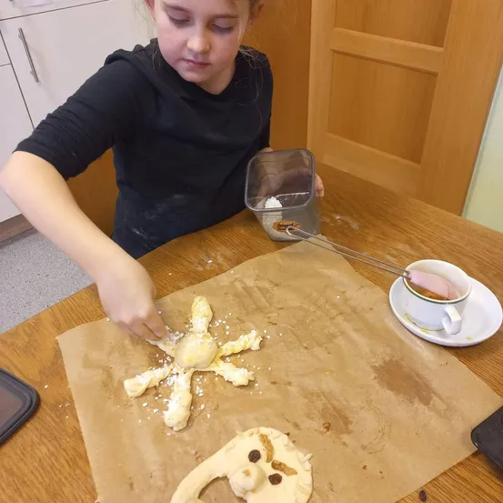 Ein junges Mädchen sitzt an einem Tisch und stellt einen dekorativen Teig her. Sie hält einen Behälter mit einem Löffel und streut etwas darauf. In der Nähe steht eine Tasse auf einer Untertasse.