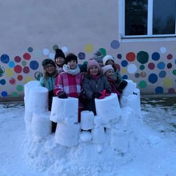 Mehrere Kinder sitzen auf einer Schneefestung im Freien. Sie lächeln und tragen warme Kleidung. Dahinter ist eine bunte Wand mit Punkten.
