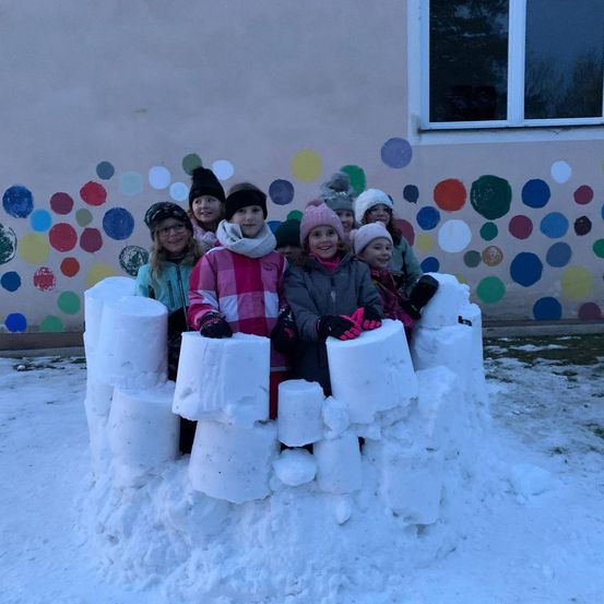 Mehrere Kinder sitzen auf einer Schneefestung im Freien. Sie lächeln und tragen warme Kleidung. Dahinter ist eine bunte Wand mit Punkten.