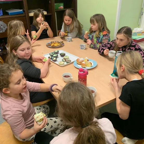 Eine Gruppe junger Mädchen sitzt um einen Tisch herum und isst Cupcakes und Donuts, vor ihnen stehen Tassen und Teller.