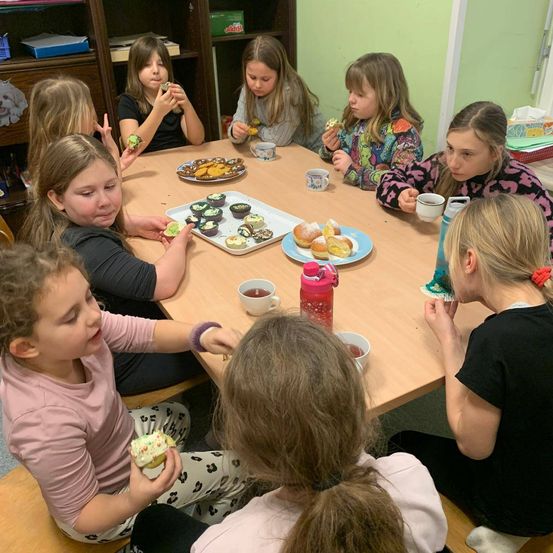 Eine Gruppe junger Mädchen sitzt um einen Tisch herum und isst Cupcakes und Donuts, vor ihnen stehen Tassen und Teller.