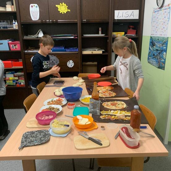 Zwei Kinder bereiten in einem Klassenzimmer Pizza mit verschiedenen Belägen auf dem Tisch zu.