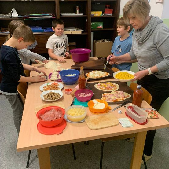 Mehrere Kinder und eine erwachsene Frau bereiten Pizza zu. Eine Frau bereitet eine Schüssel Mais vor.