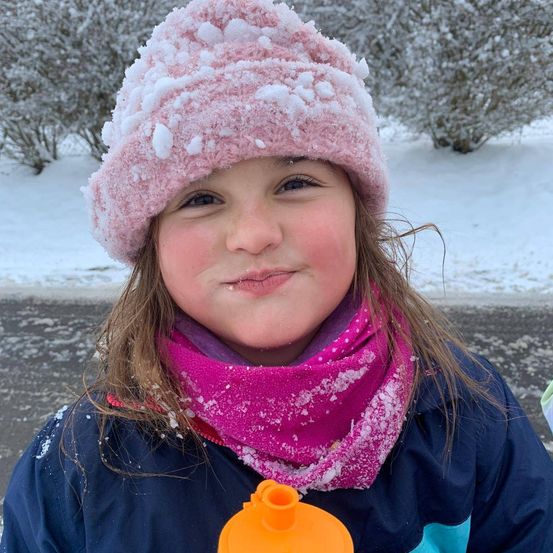 Ein junges Mädchen mit rosa Mütze und Schal lächelt im Schnee und steht draußen an einem Wintertag.