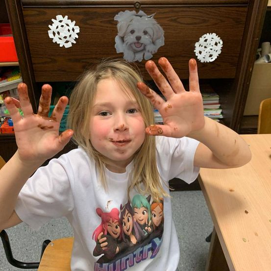 Ein junges Mädchen mit schokoladenbedeckten Händen sitzt an einem Schreibtisch und lächelt. Hinter ihr zeigt ein Holzschrank Schneeflocken und einen Hundeaufkleber.