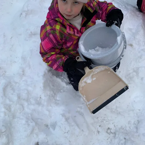 Ein kleines Mädchen in einer pinken karierten Jacke schaufelt Schnee mit einem kleinen Schneeschieber. Sie hält einen grauen Eimer voller Schnee und steht in einer verschneiten Umgebung.