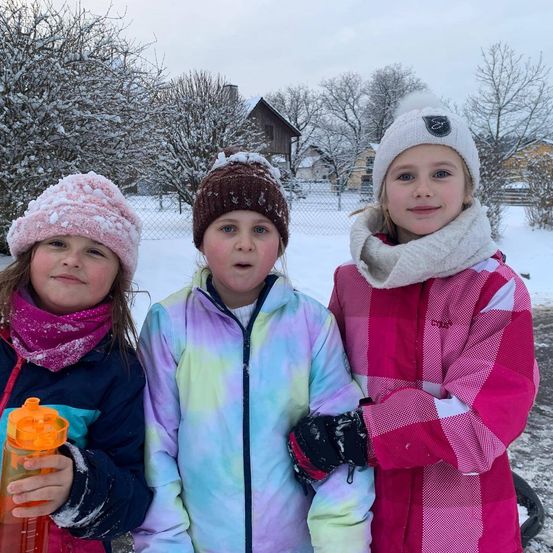Drei junge Mädchen stehen draußen im Schnee, alle in Winterkleidung. Eines hat eine rosa Mütze, ein anderes ein Tie-Dye-Jacke, und das dritte ein rosa kariertes Kostüm.