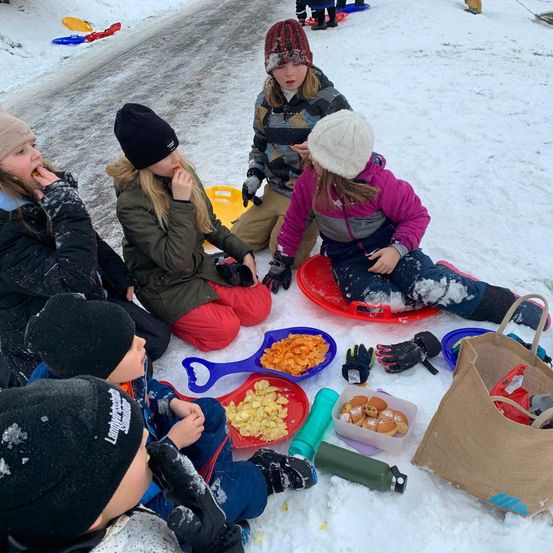 Eine Gruppe von Kindern sitzt auf Schlitten im Schnee, isst und spielt. Einige haben Mützen und Handschuhe an. Vor ihnen befinden sich Essensplatten, Handschuhe und eine Tasche. Hinter ihnen liegen weitere Schlitten im Schnee.