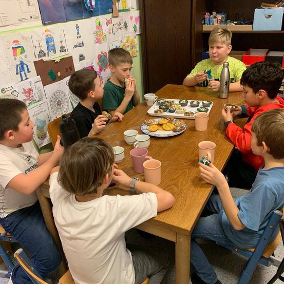 Eine Gruppe von Jungen sitzt um einen Tisch herum, isst Kekse und trinkt aus rosa Bechern. Der Tisch ist mit Platten von Keksen besetzt, und ein Junge in einem gelben T-Shirt hält einen Snack.