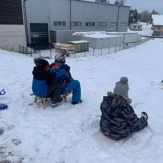 Drei Kinder sitzen im Schnee. Zwei sind auf einem hölzernen Schlitten, während das dritte im Schnee sitzt. Dahinter ist ein Zaun und ein Gebäude.