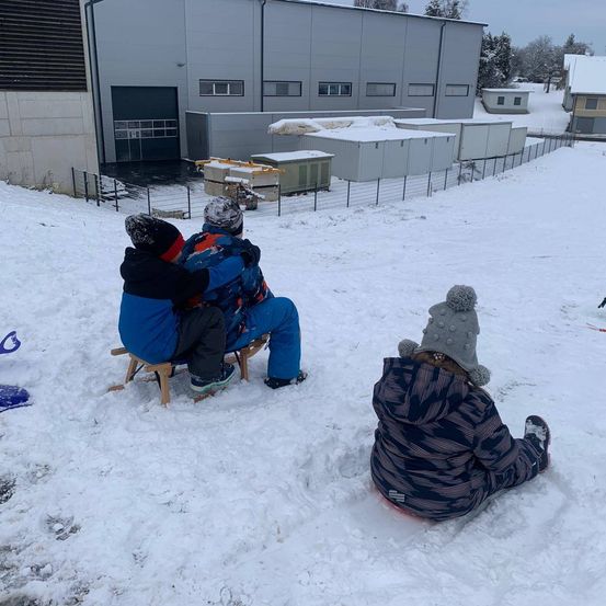 Drei Kinder sitzen im Schnee. Zwei sind auf einem hölzernen Schlitten, während das dritte im Schnee sitzt. Dahinter ist ein Zaun und ein Gebäude.
