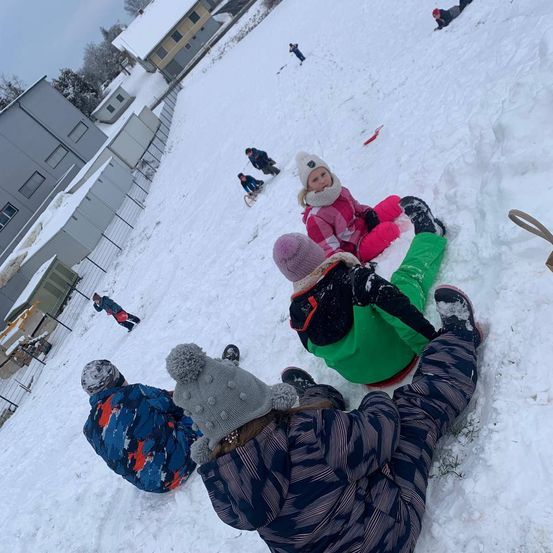 Eine Gruppe von Kindern spielt im Schnee, einige rutschen den Hang hinunter, andere sitzen auf Schlitten. Im Hintergrund sind Gebäude und ein Zaun zu sehen.