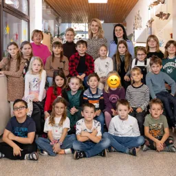 Eine Gruppe von Kindern und Erwachsenen befindet sich in einem Raum, einige sitzen und andere stehen. Sie lächeln und eine Frau lächelt in die Kamera.