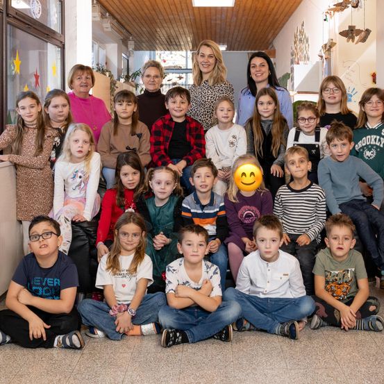 Eine Gruppe von Kindern und Erwachsenen befindet sich in einem Raum, einige sitzen und andere stehen. Sie lächeln und eine Frau lächelt in die Kamera.