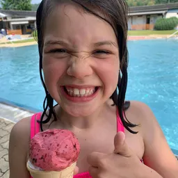 Ein junges Mädchen mit nassen Haaren lächelt und zeigt einen Daumen hoch, während es einen Erdbeereisbecher hält. Sie steht in der Nähe eines Pools.