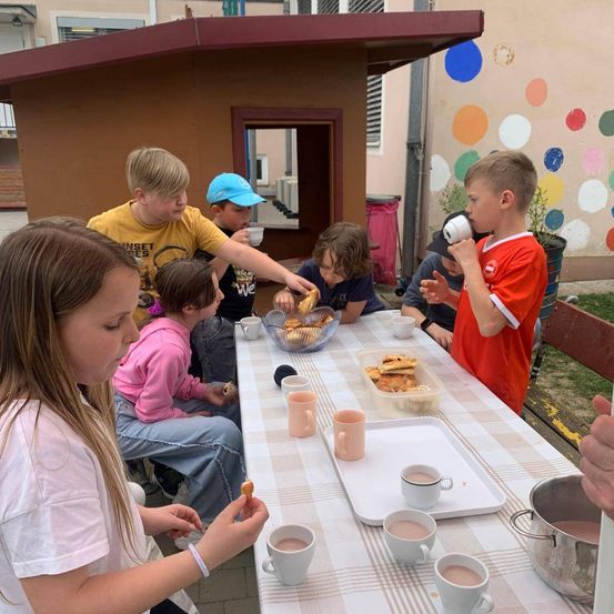 Mehrere Kinder sitzen draußen an einem Tisch mit Essen und Getränken, ein Junge trinkt aus einem Becher. Auf dem Tisch stehen Tassen, eine Schüssel mit Essen und ein Topf. Die Wand hinter ihnen hat bunte Kreise.