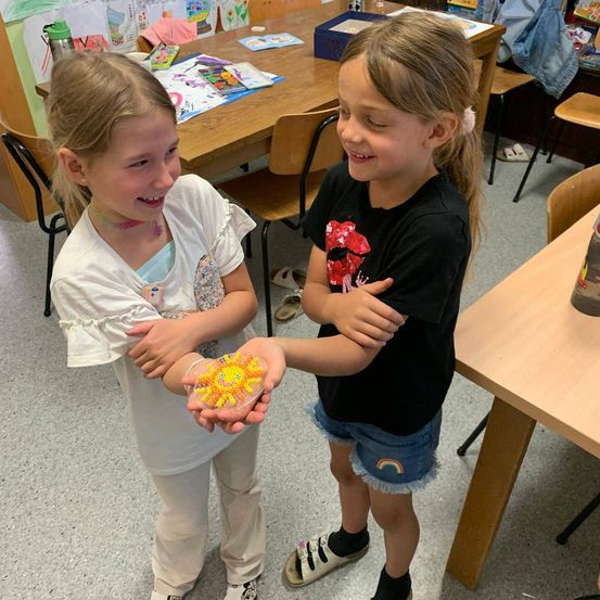 Zwei junge Mädchen stehen in einem Klassenzimmer. Ein Mädchen hält ein kleines, gelbes Objekt in einem durchsichtigen Behälter. Das andere Mädchen lächelt und sieht es an.