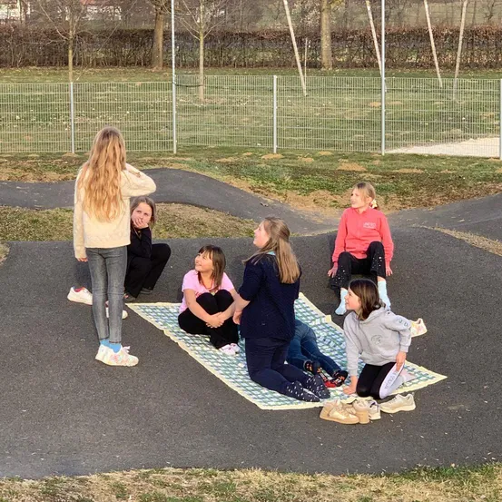 Mehrere Mädchen sitzen auf einer Decke in einem Park, zwei mit den Händen auf den Knien und eine mit den Händen am Kinn.