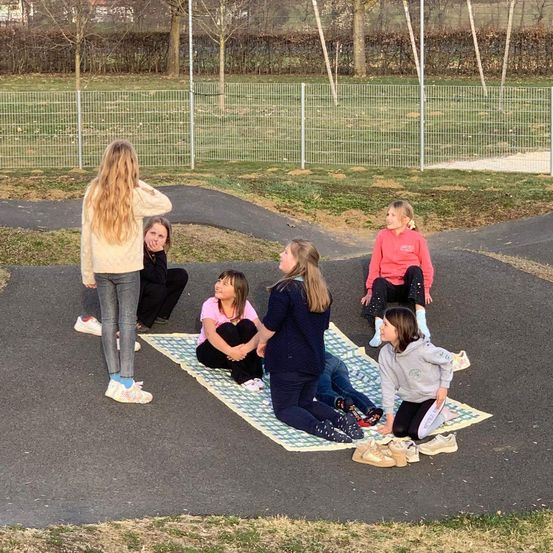 Mehrere Mädchen sitzen auf einer Decke in einem Park, zwei mit den Händen auf den Knien und eine mit den Händen am Kinn.
